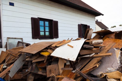 A large pile of mixed construction and demolition waste, including wooden planks, panels, and other debris, is stacked against the exterior wall of a white residential building with dark brown window shutters. The debris appears weathered and unevenly arranged, covering part of a paved driveway or pathway in a residential area, possibly in Uxbridge. The building's facade is visible in the background, with one window partially open and another closed. The scene is outdoors during daytime, with natural light highlighting the textures and colors of the wooden and materials waste. The image illustrates a typical scenario of rubbish or waste accumulation that a professional rubbish removal service, such as Office Clearance Uxbridge, might handle during an office or property clearance in the local postcode area. The waste includes various wooden items and paneling, indicating renovation or renovation debris that requires proper disposal and clearance.