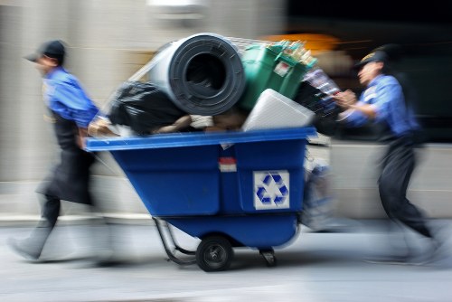 A moving blue wheeled rubbish bin filled with assorted waste items, including a large black plastic bag, a green outdoor trash bin, a white foam container, and other debris, is being pushed along a street by two individuals dressed in blue work uniforms and caps. The background is blurred, suggesting motion, and the scene appears to be part of a rubbish removal service, possibly associated with Office Clearance Uxbridge, operating within the local area of Uxbridge or the nearby postcode district. The bin is situated outdoors on a paved surface with the environment indicating urban or commercial surroundings, emphasizing the waste collection or clearance activity as part of professional rubbish disposal services.