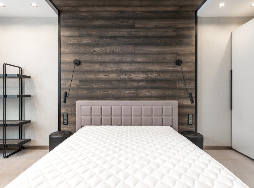 A bedroom interior featuring a large, light-colored upholstered bed with a thick, tufted white mattress positioned centrally against a beige wall. On either side of the bed, there are matching dark wo