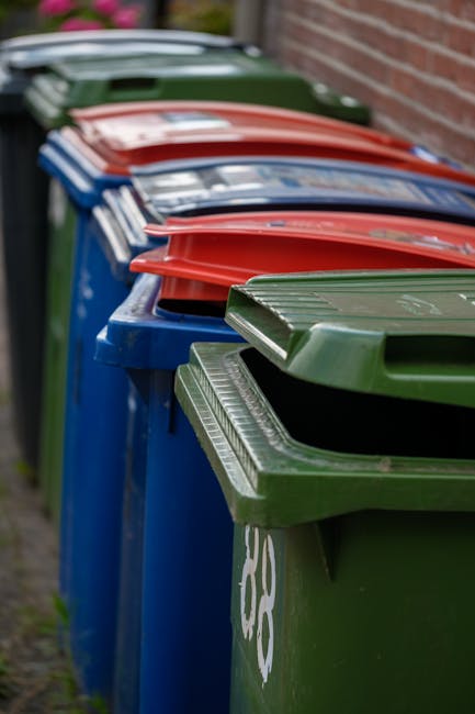 A tall, bright blue wheeled rubbish bin with a closed lid, positioned outdoors on a grassy area near a paved surface. The bin has a yellow and blue sticker with the text 'FIGHT BACK!' attached to its