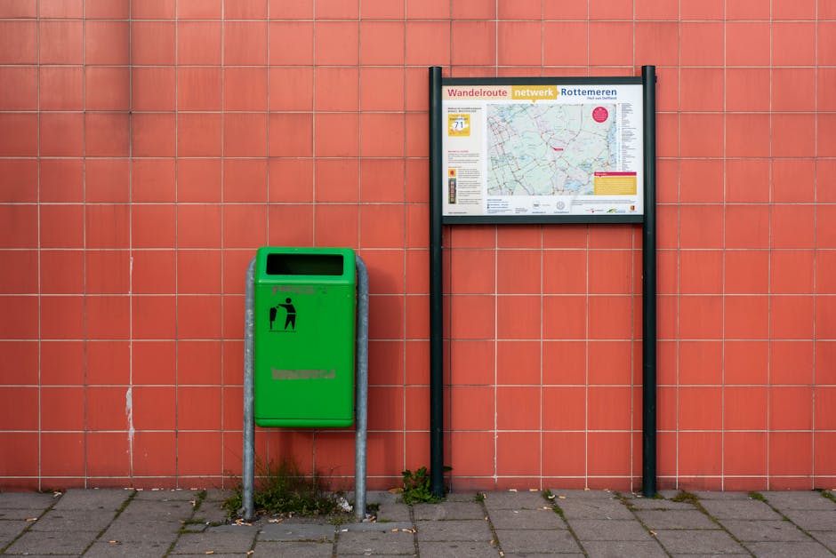The image depicts a green public waste or recycling bin mounted on a metal stand situated against a red tiled wall. The bin has a black open slot on top for depositing waste and features a black picto