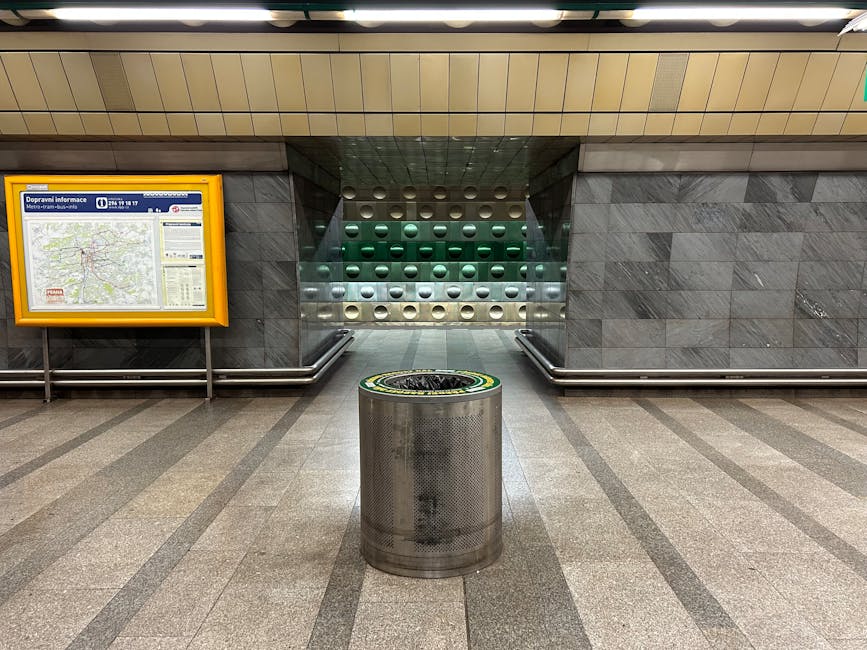 The image depicts a green public waste or recycling bin mounted on a metal stand situated against a red tiled wall. The bin has a black open slot on top for depositing waste and features a black picto
