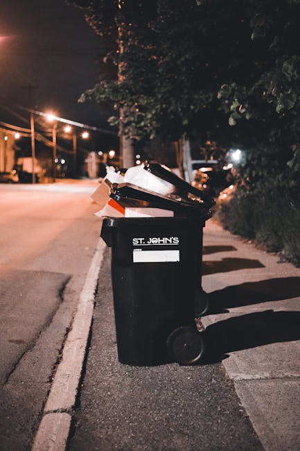 A busy urban street scene showing a pedestrian walking away from the camera, carrying a green bag of rubbish, with several other pedestrians visible on the sidewalks. The street is lined with small sh